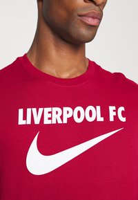 Punainen puuvillainen T-paita, jossa on valkoinen "LIVERPOOL FC" -teksti ja valkoinen Nike-swoosh-logo. Pyöreä kaula-aukko ja lyhyet hihat.