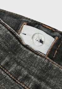 Tessuto denim nero con un motivo distorto, caratterizzato da un'etichetta bianca e un bottone trasparente. Cuciture marroni visibili in evidenza. Vista ravvicinata.