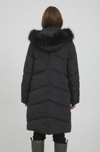 Sort pufferjakke med chevronmønster, en hætte med faux fur-fôr og en mellemlang længde, båret over sorte bukser og grønne støvler.