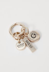 Portachiavi in metallo con diversi charm: un charm circolare con "GUESS EST. 1981," un charm rettangolare con "GUESS," e un emblema circolare, tutti in tonalità dorate.