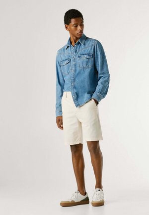 Mann som står med hendene i lommene, iført lys blå denimskjorte, hvite shorts og hvite joggesko foran ensfarget bakgrunn.