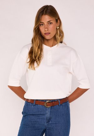 Donna con capelli lunghi e mossi, che indossa una camicia bianca con maniche a gomito abbottonata, jeans blu e una cintura marrone con fibbia dorata.