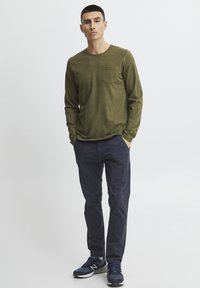 Olive green long-sleeve shirt with a chest pocket, paired with navy blue pants and navy sneakers. Smooth fabric, relaxed fit, minimalistic design.