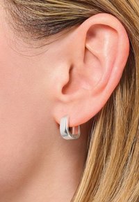 Primo piano di un orecchio femminile con capelli biondi lisci, che indossa un orecchino a cerchio spesso in argento lucido.