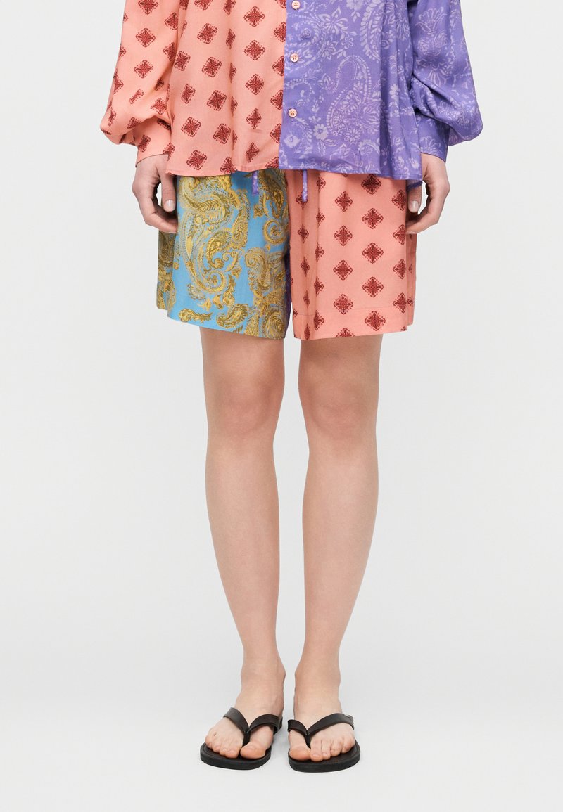 Osoba nosiaca šortky s nezhodnými vzormi a polorukávové tričko, v čiernych žabkách, stojaca na bielom pozadí.