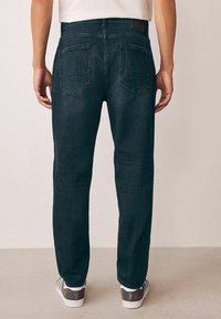 Jeans din denim de culoare bleu închis, cu croială dreaptă, două buzunare la spate și detalii de cusătură contrastante. Purtate cu adidași gri.