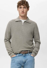 Maglione a maglia grigio chiaro con zip e colletto, caratterizzato da un motivo strutturato, abbinato a jeans azzurri slavati e cintura marrone.