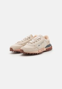 Beige színű sportcipők, amelyek textúrázott felsőrésszel, velúr részletekkel és kiemelkedő mintázatú gumitalppal rendelkeznek. A cipő oldalán Lacoste márkajelzés található.
