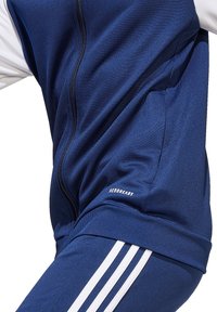 Blauwe sportieve trainingspak gemaakt van gestructureerde stof met ritsluiting, featuring witte strepen op de mouwen en een klein "AEROREADY" logo detail.