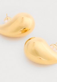 Náušnice zlatého odtieňa s hladkým tvarom slzy a lesklým povrchom; majú priesvitné podloženie pre bezpečné nosenie.