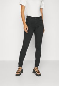 Mujer que lleva unos pantalones vaqueros ajustados negros combinados con sandalias de tacón cuadrado negras y una blusa blanca de manga corta, de pie contra un fondo liso.