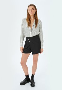 Camisa de botones a rayas en blanco y negro con un escote en V profundo, combinada con pantalones cortos negros con un diseño envolvente y dos botones plateados.
