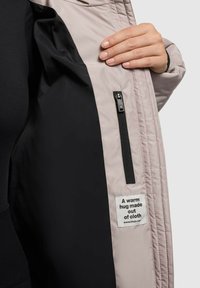 Leichte, beigefarbene Jacke mit schwarzem Innenfutter, ausgestattet mit einer Reißverschlusstasche und einem Etikett mit der Aufschrift "Eine warme Umarmung aus Stoff."