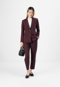 Kvinne iført en mørk lilla skreddersydd dress med en hvit skjorte, som holder en svart håndveske og har på seg mørk lilla loafers, står foran en hvit bakgrunn.
