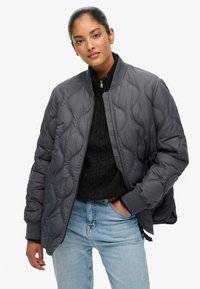 Giubbotto bomber grigio trapuntato con colletto alto, polsini a costine e tasche laterali, indossato sopra un maglione nero a trecce e jeans in denim.