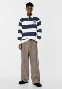Polo s dugim rukavima u tamnoplavo-bijelim prugama s ovratnikom i logom na prsima, uparen s hlačama širokih nogavica od teksturirane bež tkanine i crnim čizmama.