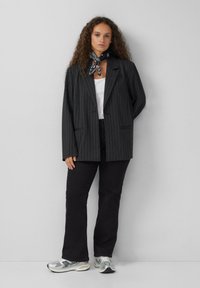 Vrouw die tegen een grijze muur staat, gekleed in een zwart gestreepte blazer, een witte top, zwarte broek, zilveren sneakers en een zwart gemusterde halsdoek.
