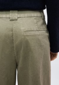 Κοντινό πλάνο ελιάς πράσινου παντελονιού με μια μόνο πίσω πόρπη και θηλιά ζώνης από ύφασμα με κεντημένο λογότυπο, φορεμένο με σκούρο πουλόβερ.