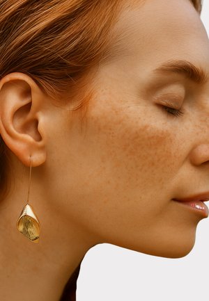 Pendientes de gota en tono dorado con un diseño en forma de hoja y una superficie texturizada, colgando de un alambre delgado. La forma general es alargada y orgánica.