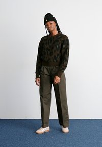 Bruin luipaardprint sweater, olijfgroene broek en beige sneakers. Model draagt een zwarte beanie van gestructureerde stof met een relaxte pasvorm.