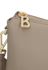 Angolo di una borsa in pelle beige con texture, dotata di una cerniera dorata con un tirante a forma di lettera "B" e un gancio dorato di attacco sulla tracolla.