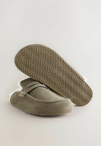 Chaussons en suède gris clair avec doublure en peluche, semelle en caoutchouc plate ornée d'un motif en chevrons, et un détail de strap décoratif.