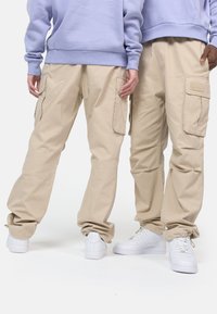 Beige cargos med flere sidelommer, avslappet passform og snøre i kante, kombinert med hvite sneakers. Lys lilla genser i bakgrunnen.