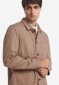 Brun tygjacka med krage, knäppning framtill och sidoficka. Slät textur med en avslappnad passform, prydd med stora bruna knappar.