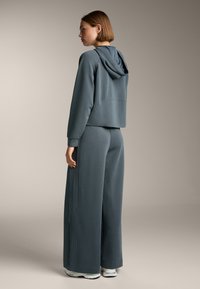 Completo a due pezzi in tessuto morbido e texturizzato. Giacca cropped con cappuccio abbinata a pantaloni larghi, entrambi in un colore blu-grigio tenue. Linee pulite.