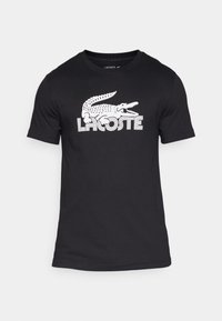 Camiseta de algodón negra con el logo del cocodrilo blanco de Lacoste en un estilo retro. Diseño de manga corta y cuello redondo.