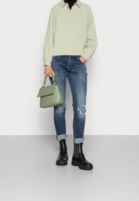 Ljust grön pullover med krage, tillsammans med slitna blå jeans och svarta ankelstövlar. Modellen håller en liten, strukturerad grön väska.