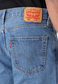 Vaaleansiniset farkut, joissa on nahkainen etiketti punaisilla grafiikoilla, takataskut sauman yksityiskohdilla sekä pieni punainen merkki sivussa.