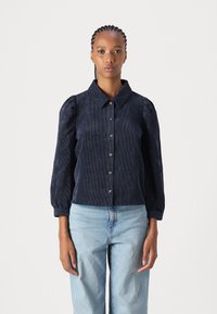 Navy corduroy shirt with a collar, button front, and puffed sleeves. Features textured vertical stripes and fitted cuffs. Paired with light jeans.