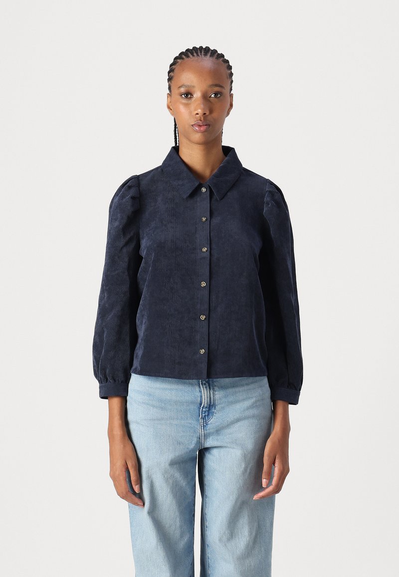 Navy corduroy shirt with a collar, button front, and puffed sleeves. Features textured vertical stripes and fitted cuffs. Paired with light jeans.
