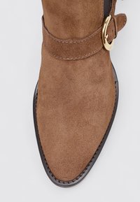 Botte de cheville en suede marron avec un bout pointu, dotée d'une semelle marron foncé cousue et d'une accroche en doré sur la sangle.