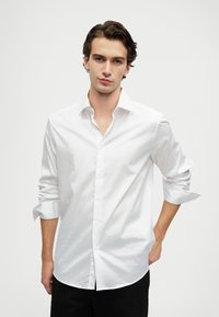 Ung man med mörkt vågigt hår som bär en vit långärmad skjorta med knappar, med uppkavlade ärmar, står med en hand i fickan.