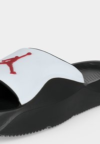 Μαύρη και λευκή σαγιονάρα slide με κόκκινο λογότυπο Jumpman στο λευκό λουράκι και υφασμένο πέλμα για καλύτερη πρόσφυση.