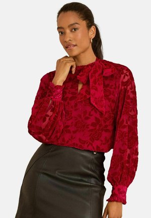 Rote Bluse mit floralem Muster, langen Puffärmeln und einem Schleifen-Halsverschluss, aus transparentem Stoff, kombiniert mit einem schwarzen Lederrock.