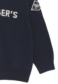 Maglione lavorato a maglia blu navy con maniche lunghe, polsini e orlo a costine. Presenta testo ricamato bianco e una toppa triangolare sulla spalla.