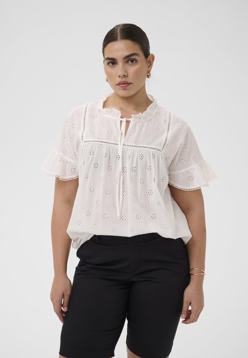 Femme portant un chemisier blanc en broderie anglaise avec des manches à volants et un short noir taille haute, debout devant un fond uni.