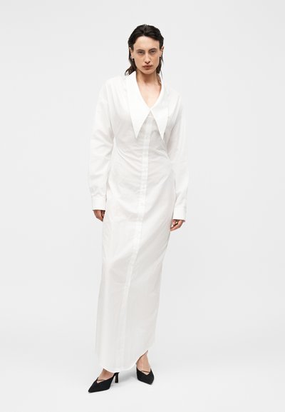 Γυναίκα με λευκό μακρύ φόρεμα με κουμπιά, φαρδύ μυτερό γιακά και μαύρες ψηλοτάκουνες γόβες, στέκεται μπροστά σε απλό λευκό φόντο.