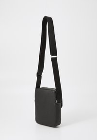 Sort læder crossbody taske med en stilren rektangulær form, justerbar vævet skulderrem og en diskret tekstureret overflade. Lynlås lukning i toppen.