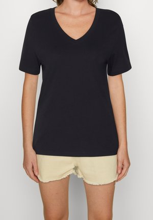 Vrouw die een effen zwart T-shirt met V-hals en korte mouwen draagt en lichtbeige gescheurde spijkershorts, staand tegen een witte achtergrond.