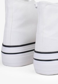 Sneakers alte bianche in tela con una parte superiore testurizzata, righe orizzontali nere sulla suola in gomma e occhielli argentati per i lacci.