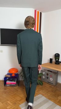 Ciemnozielony garnitur o dopasowanym kroju, zapięcie na dwa guziki i rozcięcia po bokach. Brązowe buty i pasiasty wzór na ścianie podkreślają otoczenie.