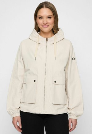 Vrouw die een lichtbeige gewatteerde jas met capuchon draagt met een voorrits, twee klepzakken en elastische manchetten, gecombineerd met een zwarte broek, glimlachend naar de camera.