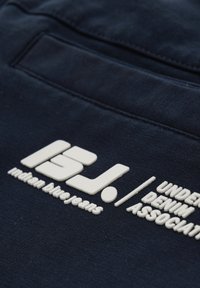 Tessuto blu navy con un logo testurizzato bianco che presenta "Indian Blue Jeans" e "Under Denim Association", posizionato sul retro.