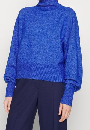 Femme portant un pull à col roulé bleu vif à manches longues et un pantalon large bleu foncé sur un fond uni.