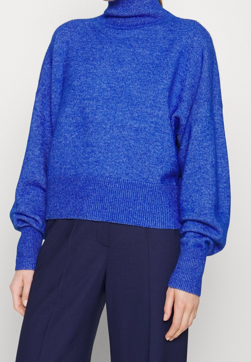 Femme portant un pull à col roulé bleu vif à manches longues et un pantalon large bleu foncé sur un fond uni.