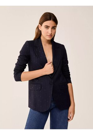 Donna con capelli castani lisci indossa un blazer blu navy a righe sottili e jeans blu, tenendo chiuso il blazer con una mano.